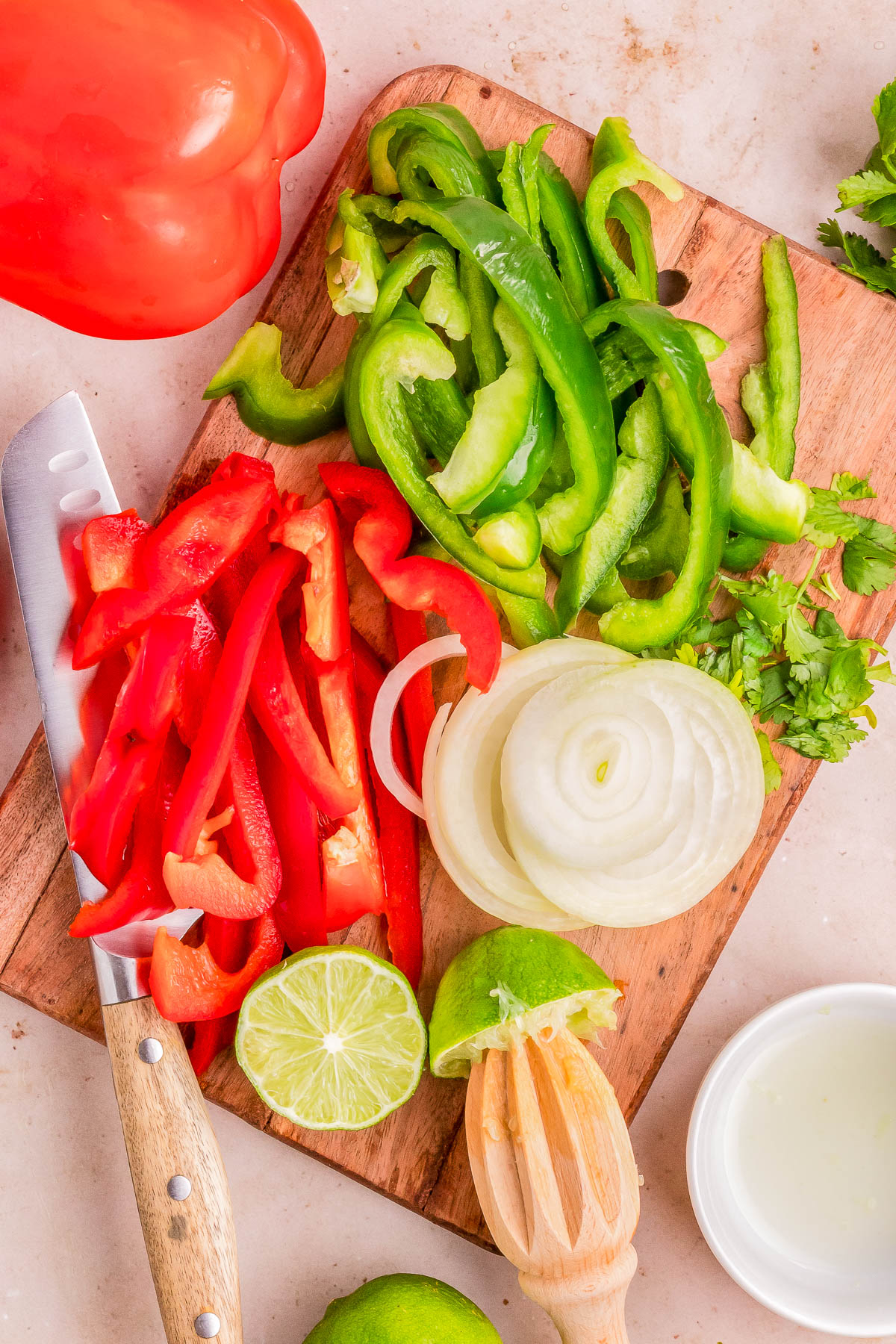 Chopped red and green bell peppers, onion, and lime on a cutting board with a knife, surrounded by whole vegetables and a wooden juicer.