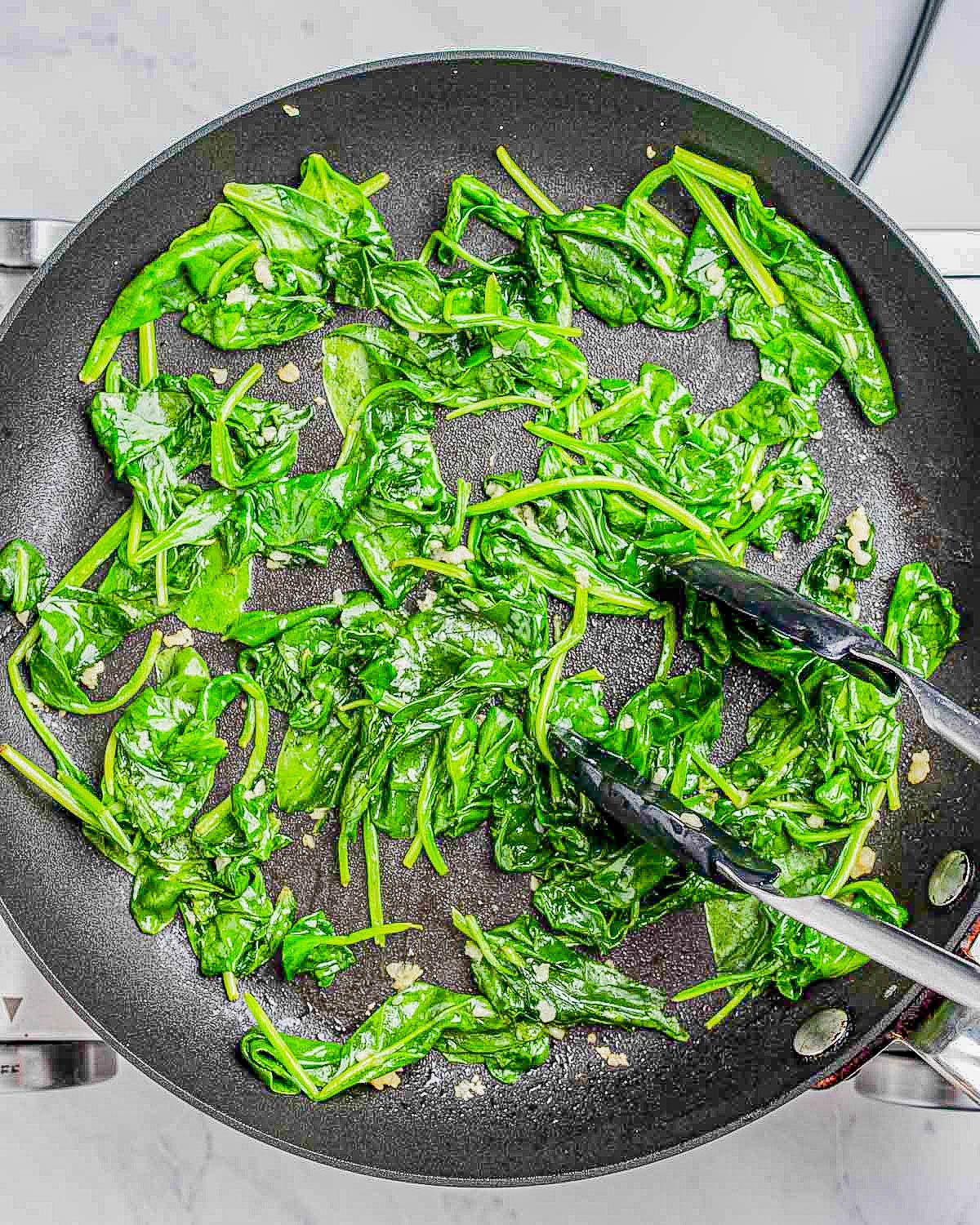 Sautéed spinach in a frying pan with tongs.