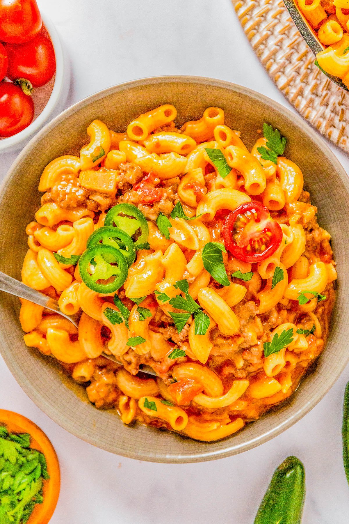 A bowl of creamy macaroni pasta topped with sliced jalapenos, cherry tomatoes, and fresh herbs, served on a white surface.