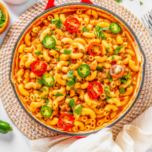 Overhead view of a pasta dish in a red skillet, topped with sliced cherry tomatoes, jalapenos, and herbs on a white surface.