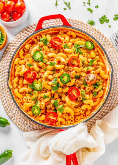 Overhead view of a pasta dish in a red skillet, topped with sliced cherry tomatoes, jalapenos, and herbs on a white surface.