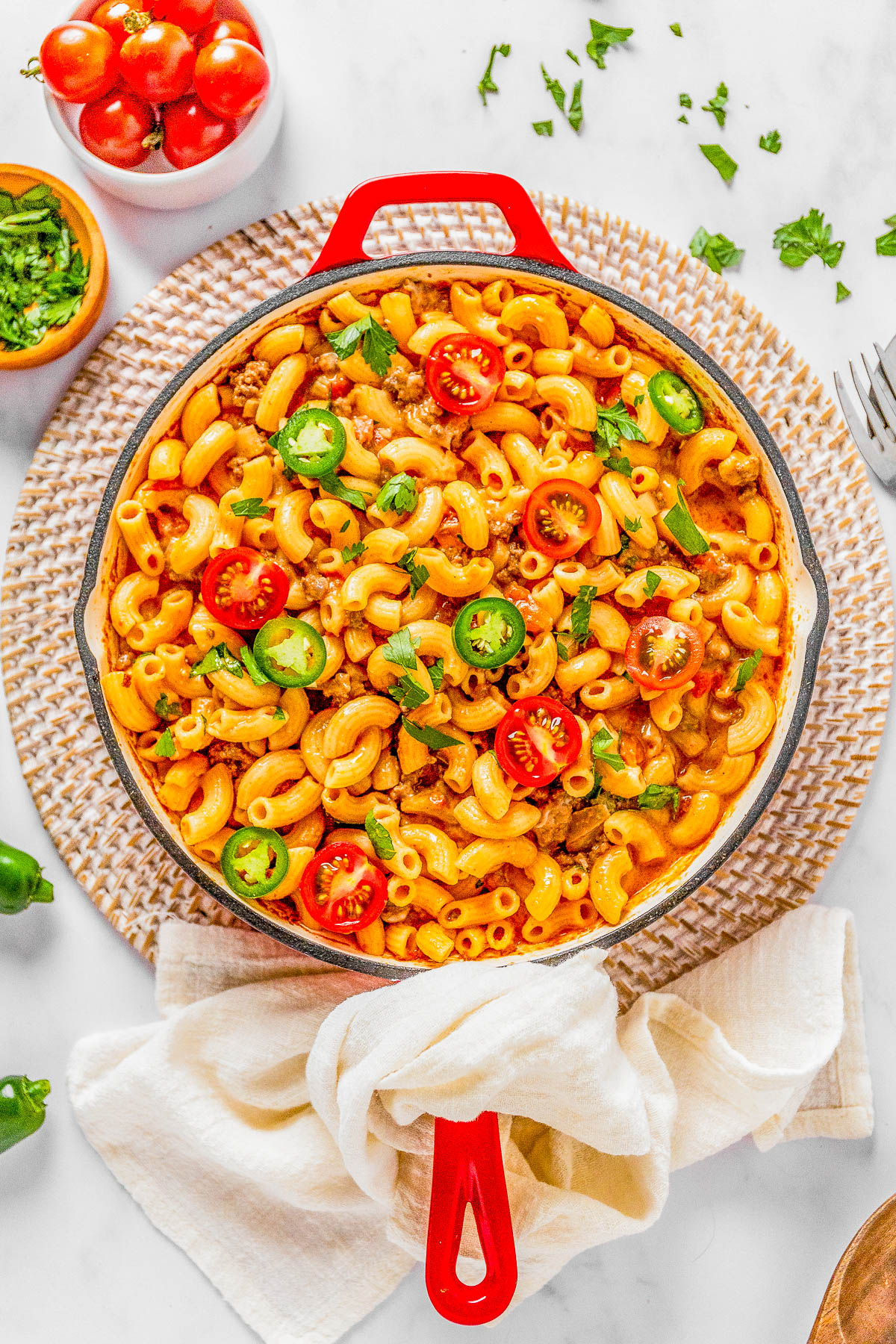 A vibrant top-down image of a red skillet filled with macaroni pasta mixed with tomatoes, green peppers, and herbs on a white background.
