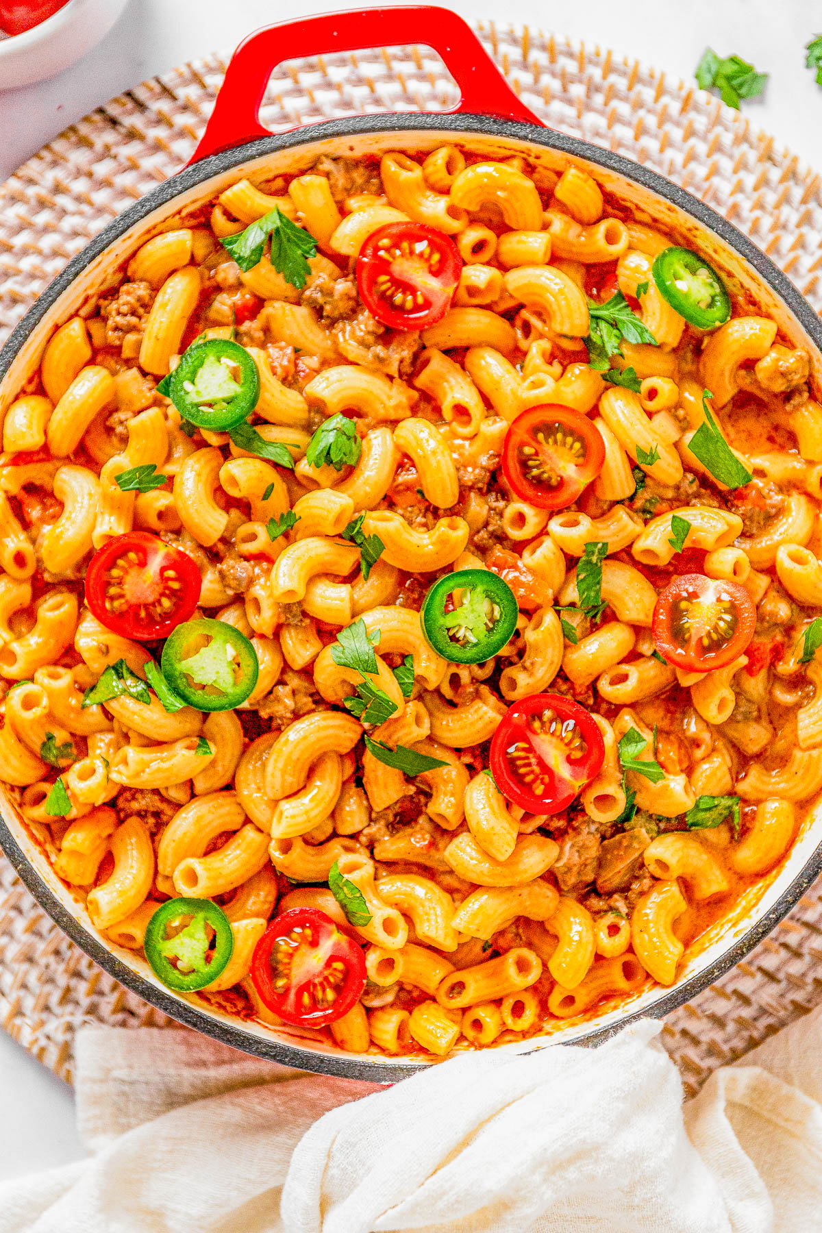 A skillet filled with macaroni pasta mixed with ground meat, sliced jalapeños, cherry tomatoes, and garnished with fresh herbs.