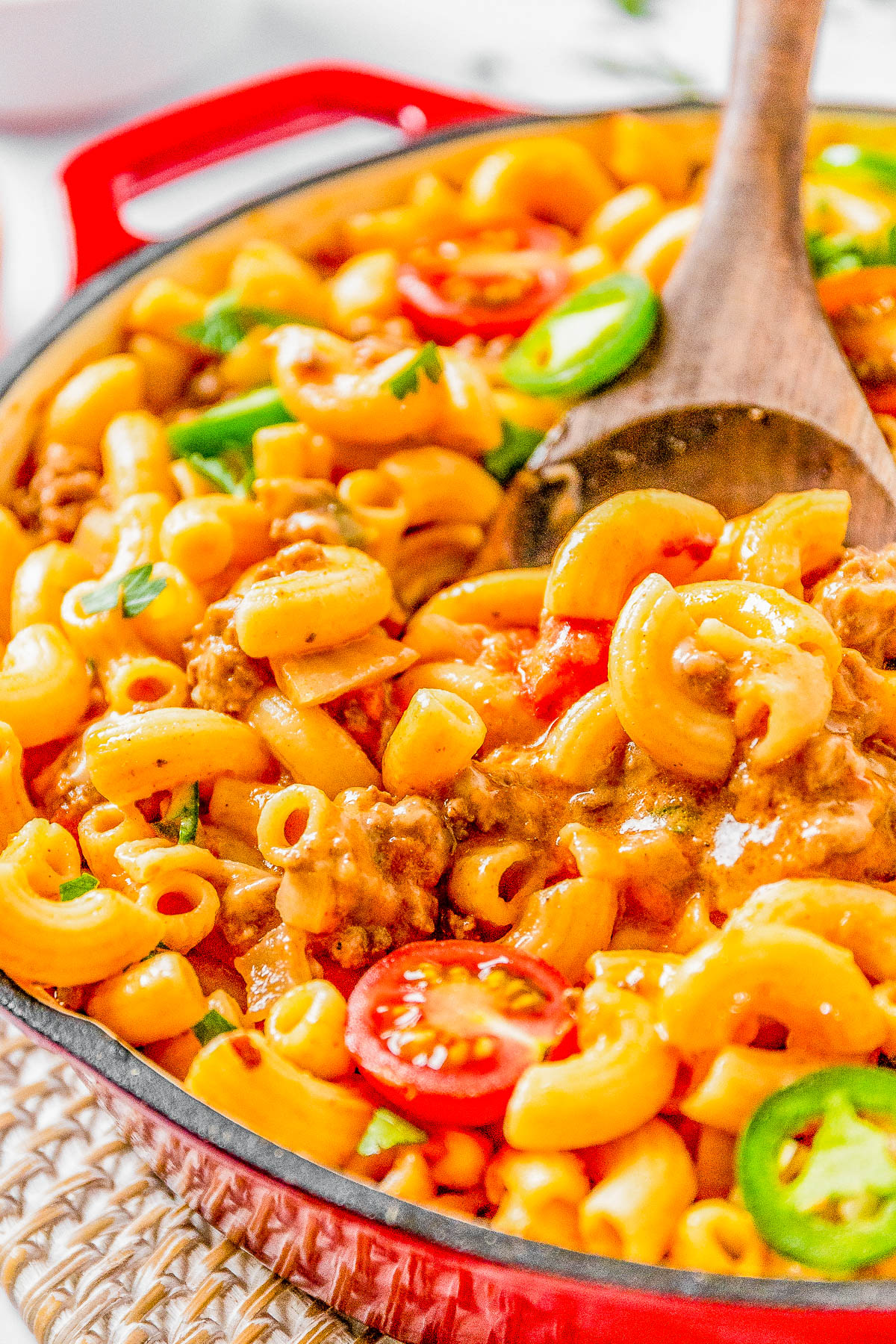 A skillet of creamy macaroni pasta with ground meat, sliced cherry tomatoes, and jalapeños, served with a wooden spoon.