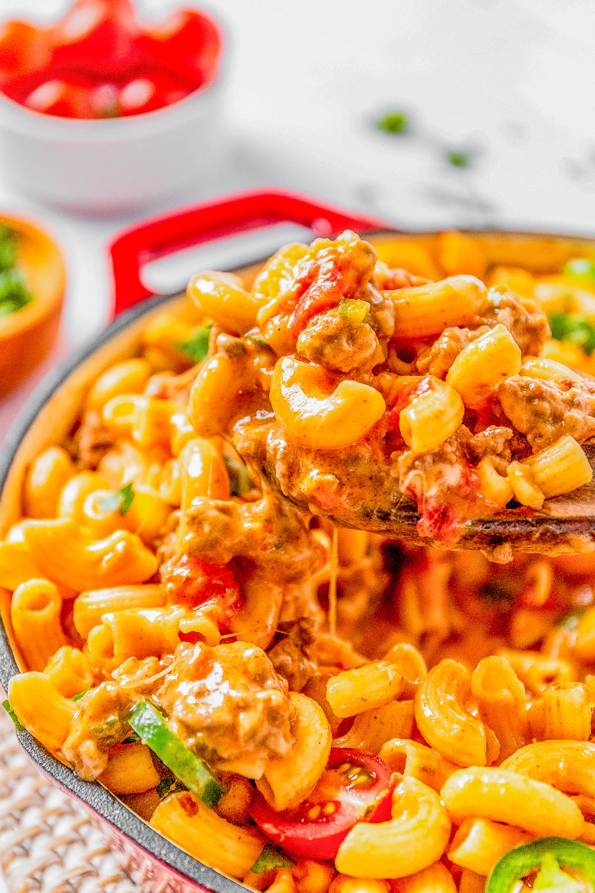 A vibrant dish of cheesy macaroni with tomatoes and minced meat in a red skillet, served on a white speckled surface.