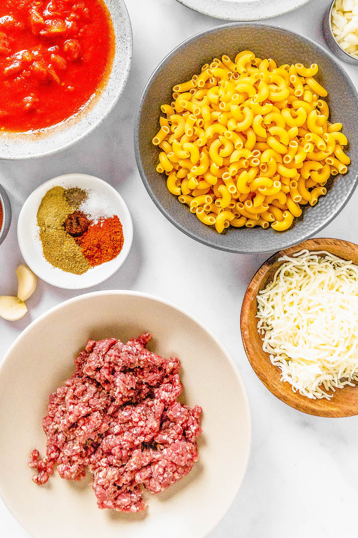 Various ingredients for cooking arranged on a table, including bowls of raw ground beef, shredded cheese, macaroni, spices, and a sauce.