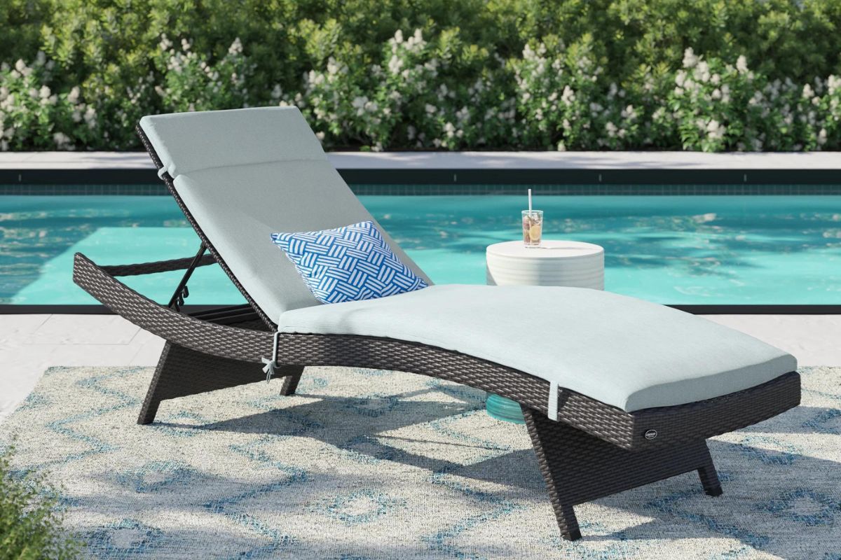 A lounge chair with a light blue cushion and decorative pillow beside a swimming pool, with a refreshing drink on a small side table.