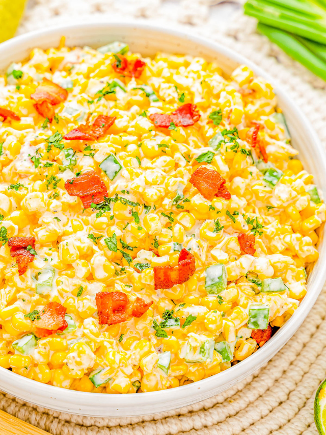 A bowl of creamy corn salad topped with chopped green onions, red peppers, and herbs.