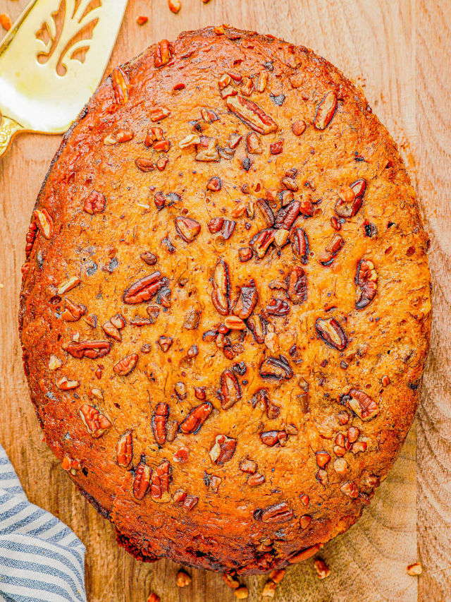 A round, baked cake with a golden-brown crust, topped with chopped pecans, placed on a wooden surface next to a brass spatula and a striped cloth.