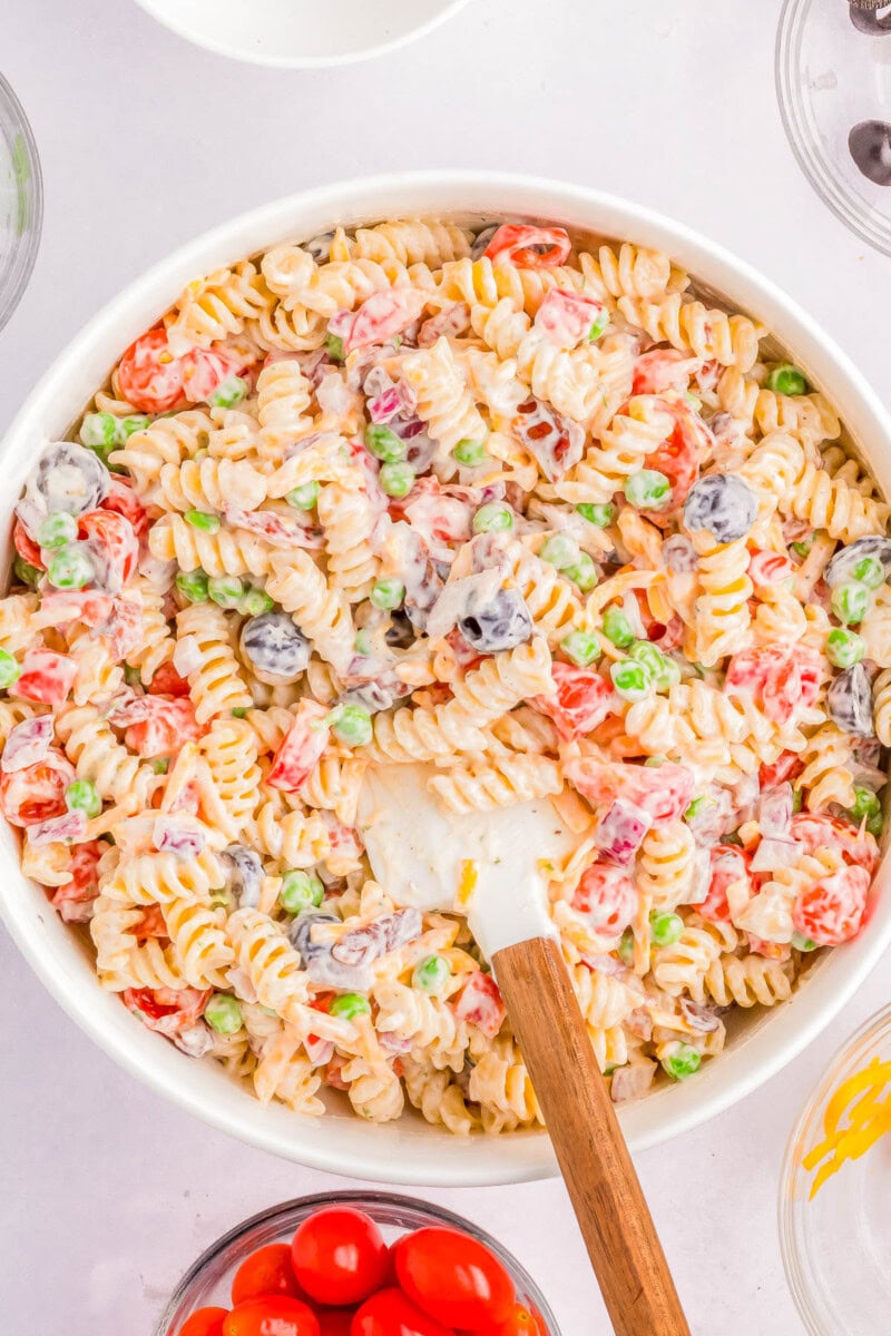 A bowl filled with creamy pasta salad containing rotini pasta, peas, cherry tomatoes, olives, and a wooden spoon for mixing.