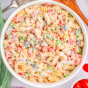 A bowl of rotini pasta salad with vegetables including peas, tomatoes, black olives, and diced red onions in a creamy dressing. Wooden serving spoons, a fork, and additional ingredients are nearby.