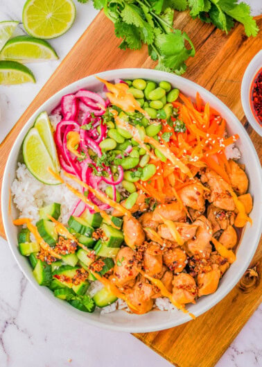 A colorful poke bowl with chicken, rice, edamame, cucumber, carrots, pickled onions, cilantro, lime, and sesame seeds, served on a wooden board.