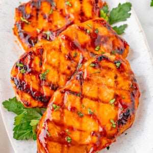Three grilled pork chops glazed with barbecue sauce are garnished with chopped herbs and arranged on a white oval plate.