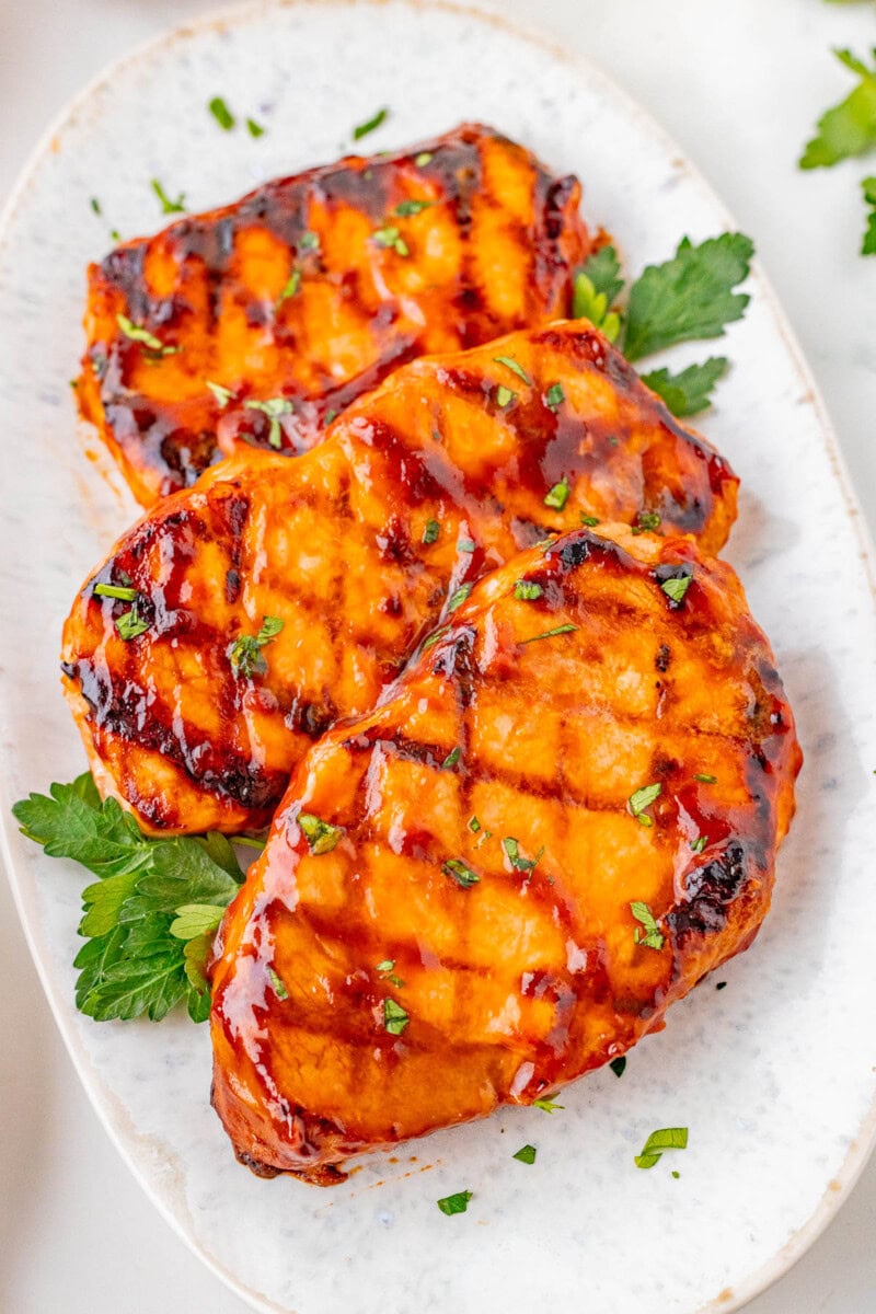 Three grilled pork chops glazed with barbecue sauce are garnished with chopped herbs and arranged on a white oval plate.