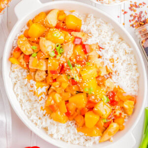 A dish of sweet and sour chicken with pineapple and bell peppers served over white rice in a white bowl. Nearby, a smaller bowl with the same dish, a red bell pepper, fresh ginger, and sesame seeds.