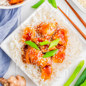 A square white plate with rice topped with glazed chicken chunks, garnished with sesame seeds and green onions. Chopsticks, fresh ginger, green onions, and a napkin are beside the plate.