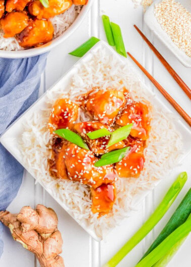A square white plate with rice topped with glazed chicken chunks, garnished with sesame seeds and green onions. Chopsticks, fresh ginger, green onions, and a napkin are beside the plate.