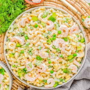 A vibrant shrimp pasta salad with green peas, dill, and cashews, served on a white platter over a rustic wicker mat and grey linen.