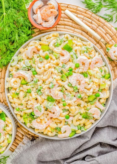 A vibrant shrimp pasta salad with green peas, dill, and cashews, served on a white platter over a rustic wicker mat and grey linen.