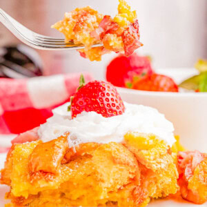 A fork holds a bite of strawberry bread pudding with a slice of cake topped with whipped cream and a strawberry on a plate below.