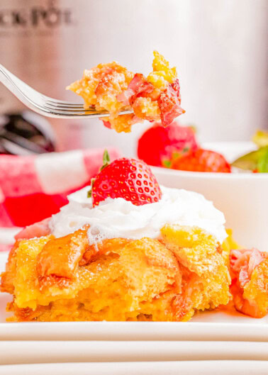 A fork holds a bite of strawberry bread pudding with a slice of cake topped with whipped cream and a strawberry on a plate below.