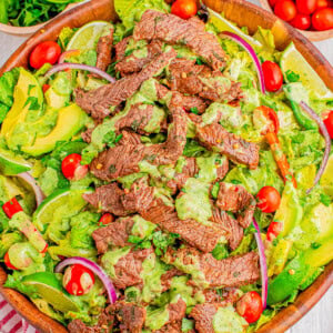 A wooden bowl filled with a salad featuring sliced steak, cherry tomatoes, avocado, red onion, and a green dressing. Other ingredients like parsley and lime are visible in the background.