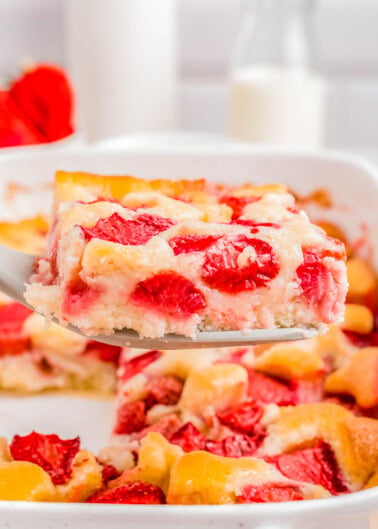 A slice of strawberry cobbler being lifted from a dish, showcasing ripe strawberries and a fluffy texture, with a glass of milk in the background.