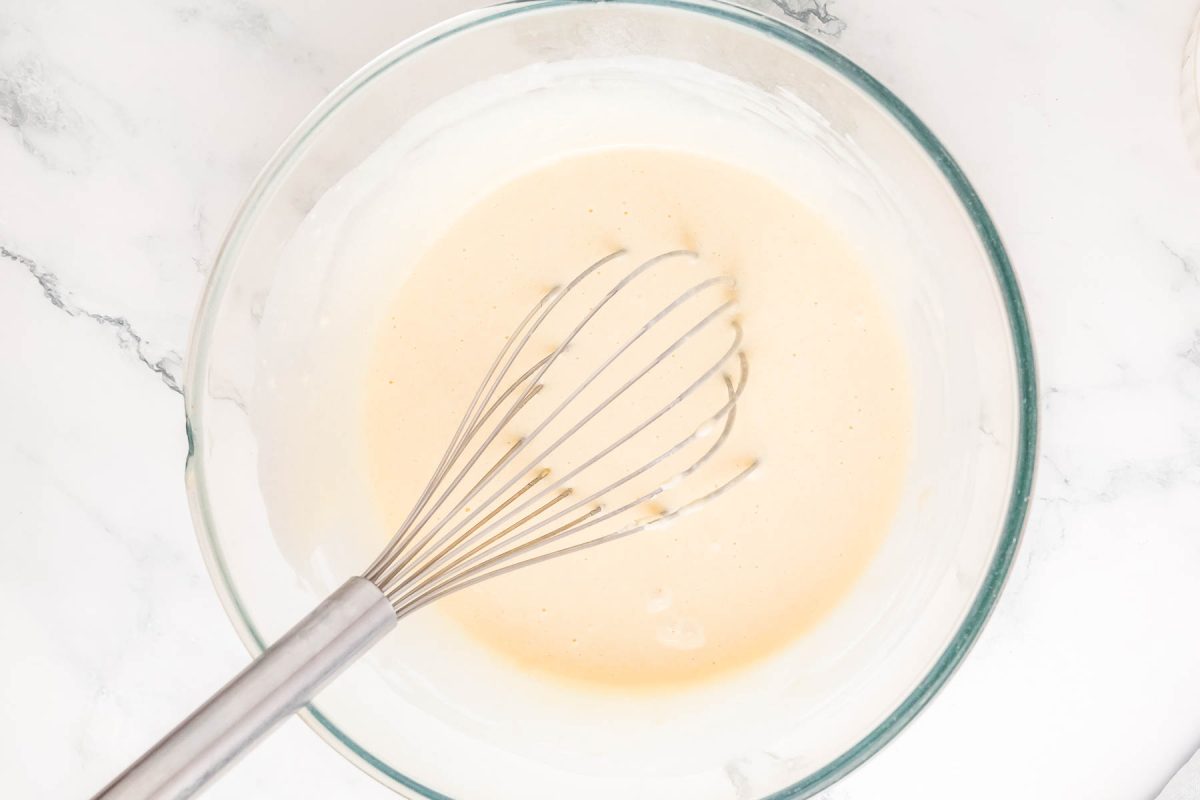 A clear glass bowl containing a light beige batter with a metal whisk, sitting on a marble surface.