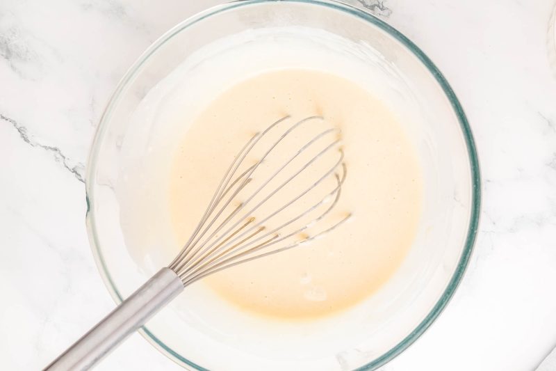 A clear glass bowl containing a light beige batter with a metal whisk, sitting on a marble surface.