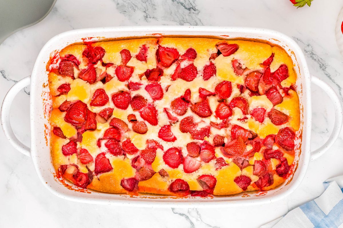 Baked dessert in a white dish with sliced peaches and strawberries on a marble surface.