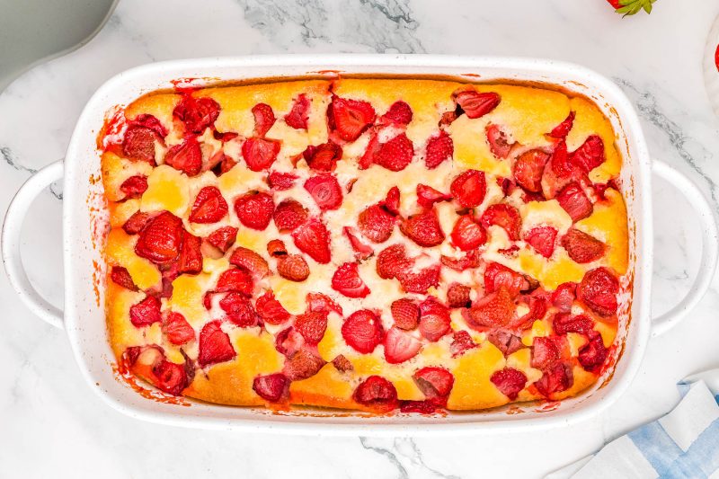 Baked dessert in a white dish with sliced peaches and strawberries on a marble surface.