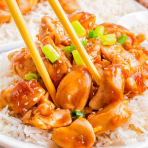 A plate of rice topped with saucy chunks of teriyaki chicken, garnished with green onions, with chopsticks picking up a piece of chicken.