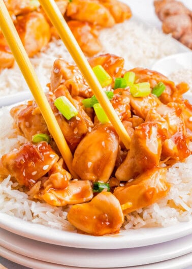 A close-up of a plate of rice topped with saucy chunks of teriyaki chicken, garnished with green onions, with chopsticks picking up a piece of chicken.