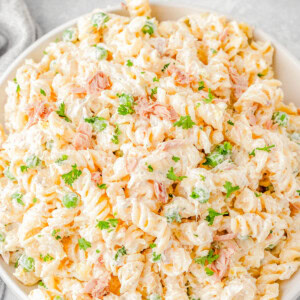 A bowl of creamy tuna pasta salad with rotini pasta, peas, and fresh parsley, placed on a light-colored surface.