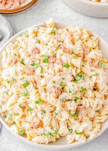 A bowl of creamy tuna pasta salad with rotini pasta, peas, and fresh parsley, placed on a light-colored surface.