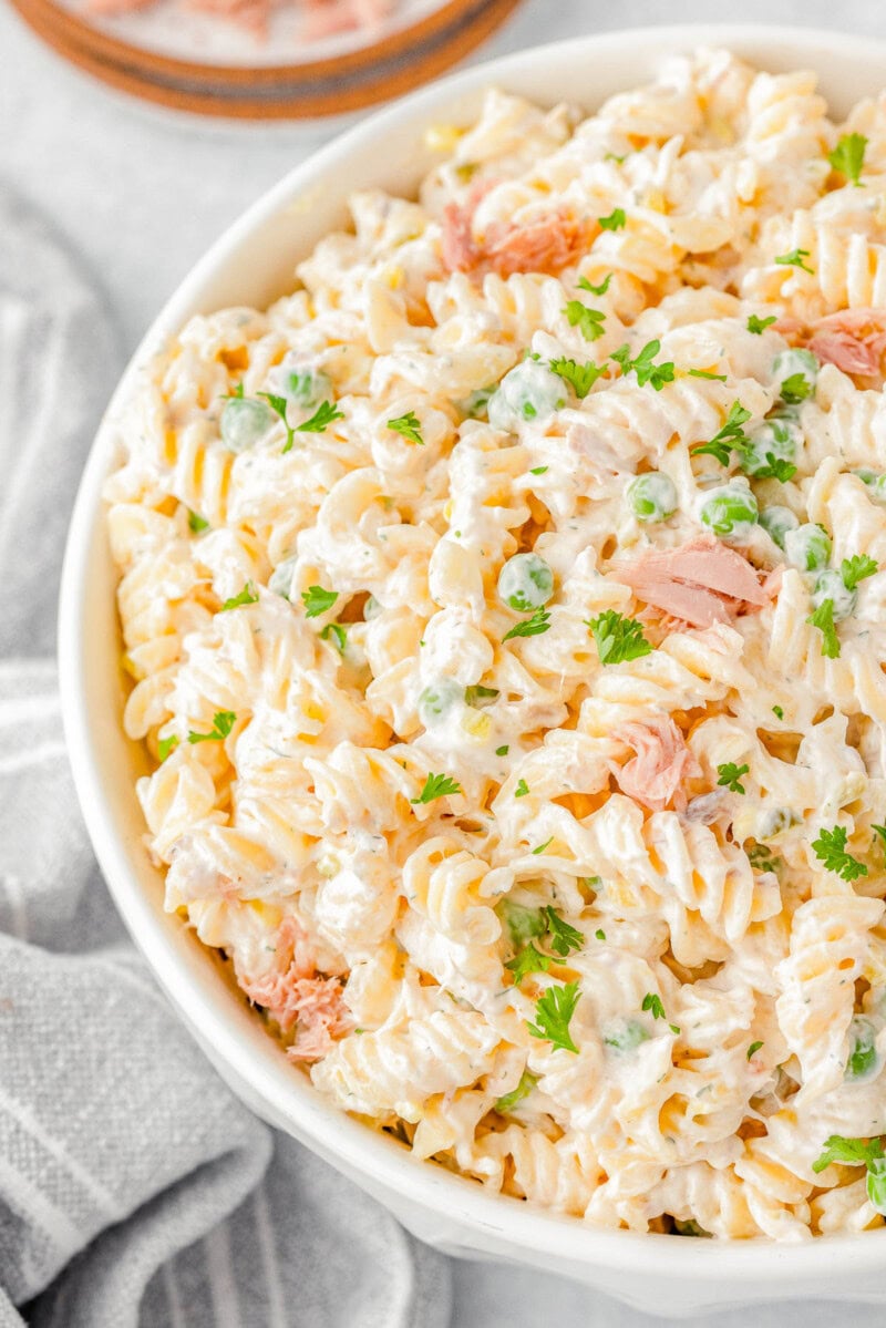 A bowl of creamy pasta salad with fusilli, peas, shredded cheese, and garnished with chopped parsley.