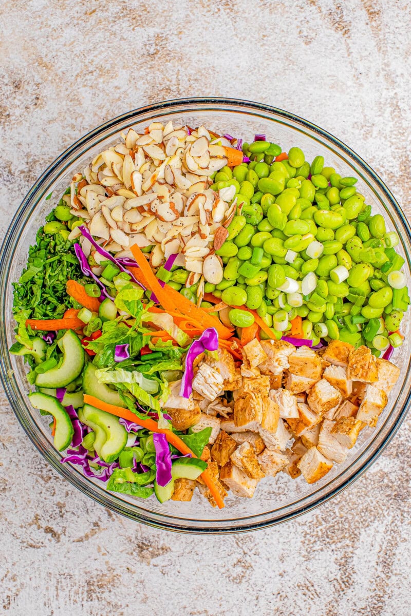 A glass bowl filled with a colorful salad consisting of sliced almonds, edamame, chopped cooked chicken, cucumber, red cabbage, carrots, and mixed greens.