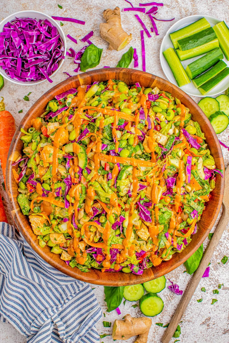 A colorful salad in a wooden bowl, featuring lettuce, purple cabbage, cucumber, carrots, edamame, and orange dressing. Plates with cucumber sticks and shredded purple cabbage are on the side. A striped cloth is nearby.