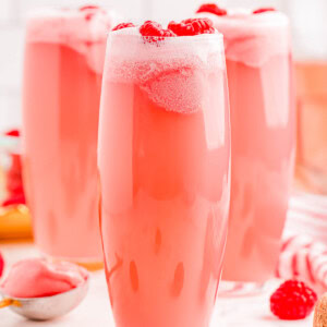 Three tall glasses filled with a frothy pink beverage, topped with raspberries, surrounded by scattered raspberries and a glimpse of a spoon with a pink substance.