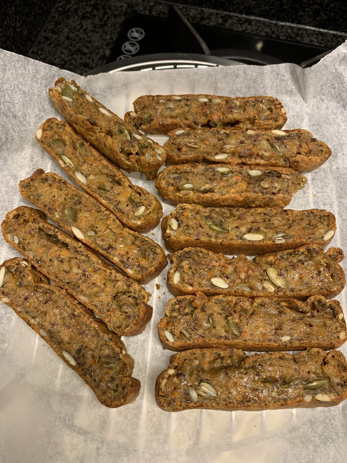 Sliced nut and seed biscotti arranged on a sheet of parchment paper, cooling on a rack in a kitchen setting.