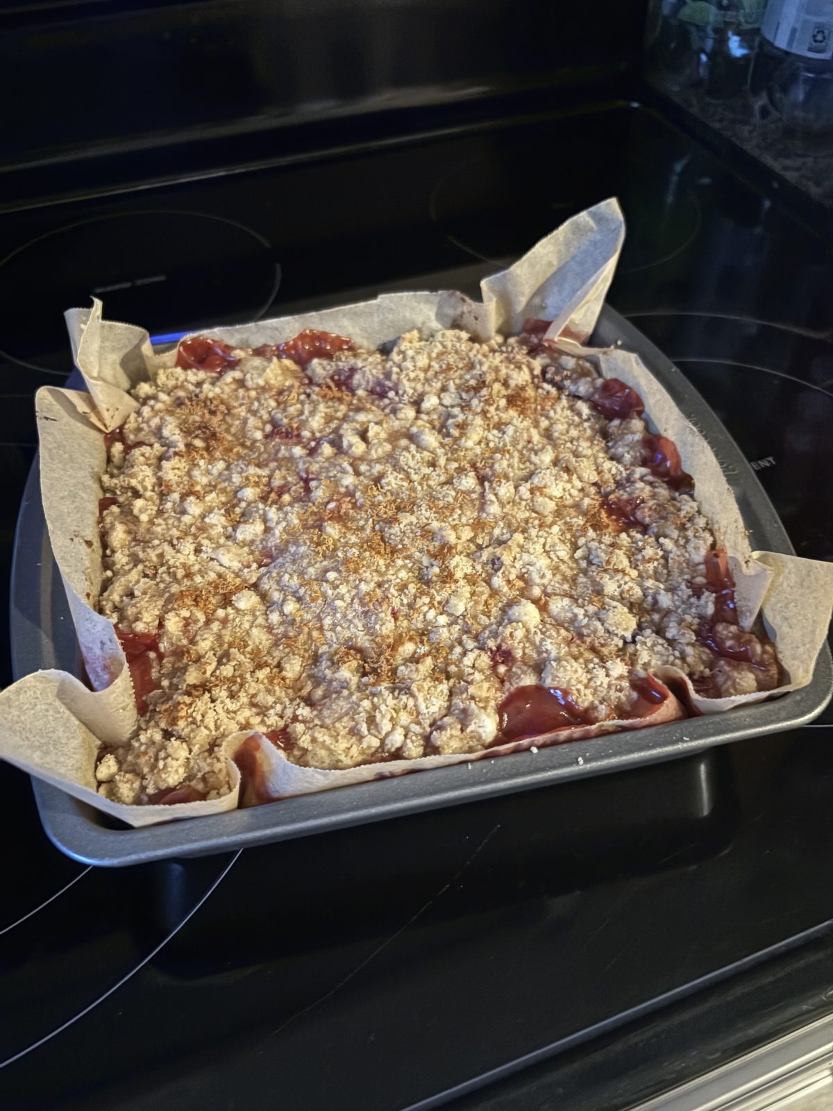 A square baking pan lined with parchment paper holds a freshly baked fruit crumble with a golden oat topping, cooling on a stovetop.