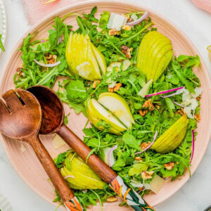 A plate of arugula salad with sliced pears, walnuts, and shaved cheese, accompanied by wooden serving utensils and gold-colored forks on a pink napkin.