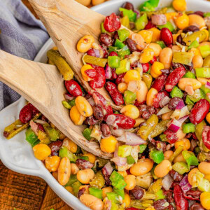 A bowl of mixed bean salad with kidney beans, black beans, corn, green bell peppers, and onions being served with wooden utensils.