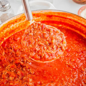 A ladle filled with hearty tomato and meat sauce hovers above a pot of the same sauce. Ingredients like parsley and salt are blurred in the background.