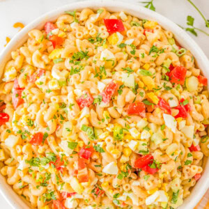 A bowl of macaroni salad with diced tomatoes, yellow bell peppers, chopped eggs, and herbs on a white surface.