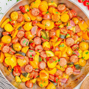 A skillet filled with cooked sausage slices, yellow bell peppers, cherry tomatoes, onions, and diced potatoes, garnished with fresh parsley, with a wooden spoon and herbs on the side.