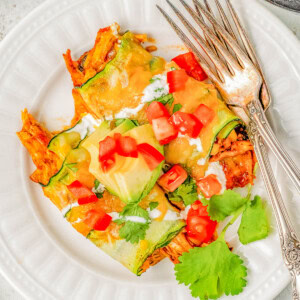 A white plate with zucchini enchiladas topped with melted cheese, diced tomatoes, avocado slices, sour cream, and cilantro. Two forks are placed on the side of the plate.