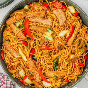 A pan of stir-fried noodles with slices of beef, red bell peppers, bok choy, and green onions. A red checkered cloth is underneath the pan. Plates and ingredients are visible in the background.