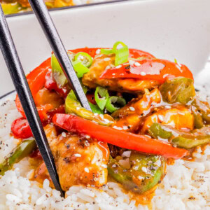 A bowl of white rice topped with stir-fried chicken, red and green bell peppers, and green onions, with a pair of black chopsticks picking up food.