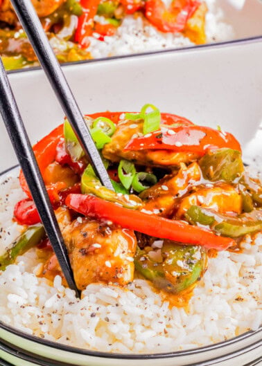 A bowl of white rice topped with stir-fried chicken, red and green bell peppers, and green onions, with a pair of black chopsticks picking up food.
